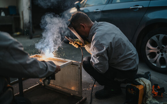 Workers in protective gear weld a metal frame outdoors beside a car, producing bright sparks and smoke. The scene captures hands-on fabrication, teamwork, and industrial craft in an outdoor setting. - Powered by Adobe