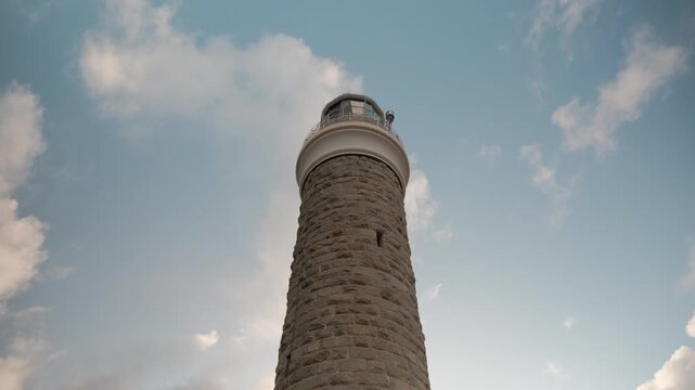 Low angle view looking up at a historic stone lighthouse tower against a blue sky with moving clouds. Majestic maritime architecture and coastal navigation beacon standing tall.