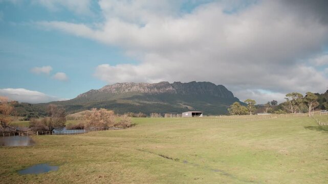 Scenic landscape of Mount Roland in Tasmania, Australia. Green farmland paddock with a rustic shed sits below the majestic rocky mountain peak under a cloudy blue sky.