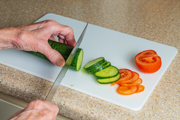 Older person preparing salad items with a cucumber and tomato on a white board