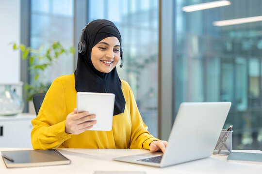 Young muslim call center agent wearing a headset and hijab, looking at a digital tablet and typing on a laptop, providing customer support or remote assistance from a modern office workspace