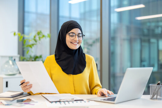 Young muslim businesswoman wearing a hijab and glasses, happily engaging with a laptop and holding a document while sitting at a modern office desk, representing professionalism and productivity - Powered by Adobe