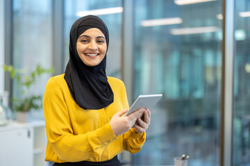Muslim businesswoman smiling at camera wearing hijab while holding a digital tablet with both hands, standing in a bright modern office against a backdrop of blurred windows