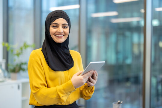 Muslim businesswoman smiling at camera wearing hijab while holding a digital tablet with both hands, standing in a bright modern office against a backdrop of blurred windows