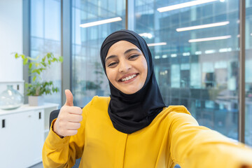 Happy young muslim businesswoman wearing a hijab and yellow blouse, smiling and giving a thumbs up gesture while taking a selfie in a contemporary corporate office environment