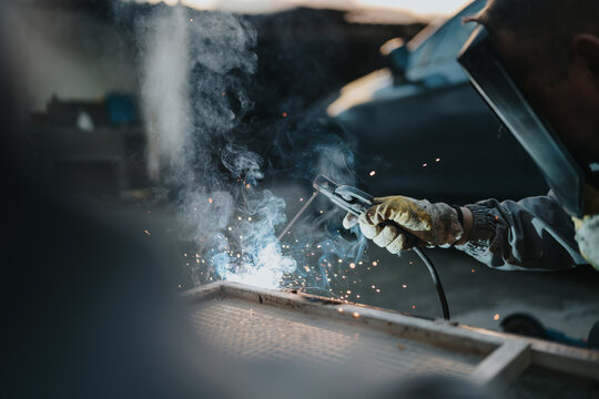 A skilled welder wearing a helmet and gloves welds metal, producing bright sparks and smoke. The scene highlights safety gear, industrial work, and focused craftsmanship in a workshop setting.
