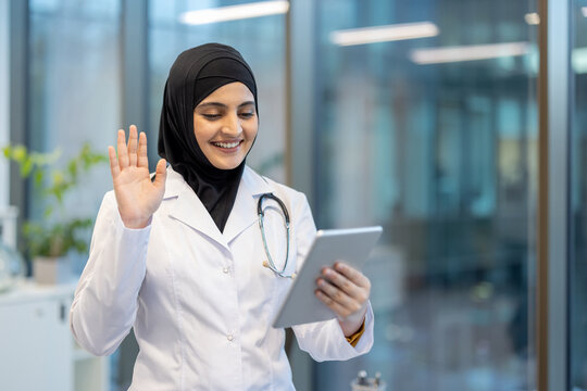 Muslim female doctor wearing a hijab and white coat smiling and waving, communicating with a patient during a virtual consultation on a digital tablet, promoting telemedicine and online healthcare