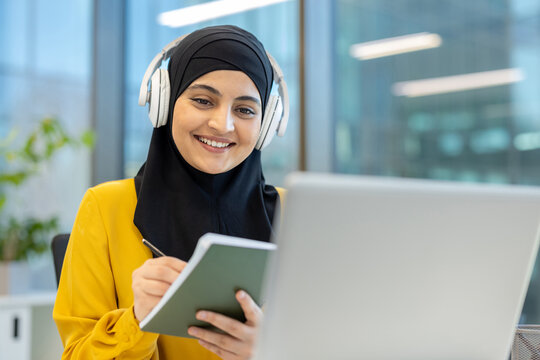 Muslim woman smiling, wearing headphones and a hijab, taking notes in a notebook while using a laptop to learn online remotely, representing e-learning, education, and career development