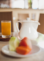 A bright orange juice glass sits next to a white teapot on a table. Blurred fruit slices are visible in the foreground.