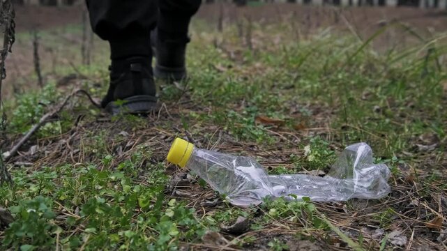 A male foot wearing a black sneaker steps firmly on a discarded plastic bottle lying in the grass. The action demonstrates the crushing of waste and the prevalence of pollution in green spaces.