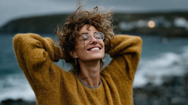 A woman with curly hair and glasses smiles broadly while standing by the shoreline, enjoying the refreshing sea breeze and the dramatic cloud-filled sky in the background. - Powered by Adobe
