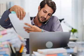 A busy businessman handles document work and takes a smartphone call in a messy office while feeling overworked