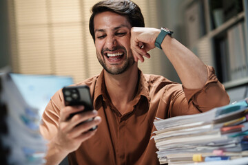A happy businessman relaxes at his desk at night, checking his smartphone after finishing work in the office.