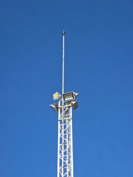 A tall, white metal lattice tower topped with high-intensity floodlights and a thin lightning rod, set against a solid, deep blue, clear sky.