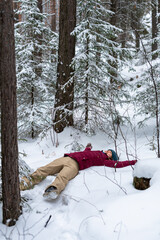 young Caucasian smiling woman lies in the snow among evergreen trees in forest She wears red jacket and beige pants, enjoying a winter day in a forested area, winter fun