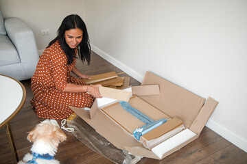 A woman is unpacking a box on the floor while a small dog watches her