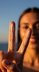 Close-up of a hand displaying the two-finger sunscreen rule with cream strips, set against a blurred smiling woman in golden sunset light, promoting skin protection and beauty care