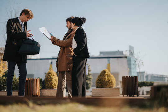 Three colleagues stand on a rooftop terrace, reviewing papers and sharing ideas. A casual business meeting in an urban setting with plants and modern buildings in the background. - Powered by Adobe