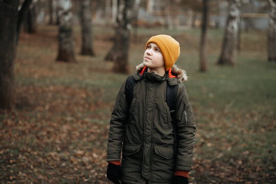 A young person in a green winter jacket and yellow knit beanie standing in an autumn park, surrounded by trees and grass.