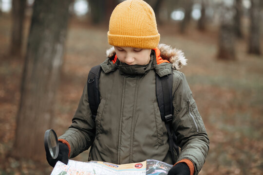 Kid teenager boy in a yellow beanie and green winter jacket studies a map while standing in an autumn forest, backpack on, ready for adventure.