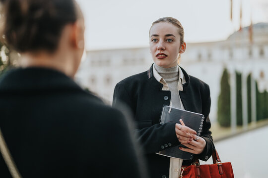 A professional young woman chats with coworkers outdoors. She wears a dark jacket, carries a notebook, and holds a red bag, suggesting collaboration and casual business discussion.