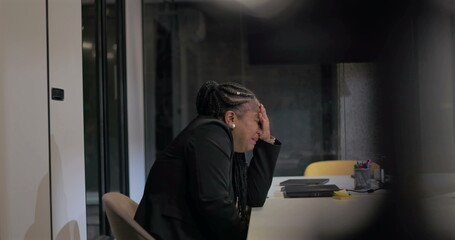 Fototapeta premium Distant view of African American corporate employee with head in hand through glass meeting room, portraying emotional burnout, fatigue, and workplace isolation