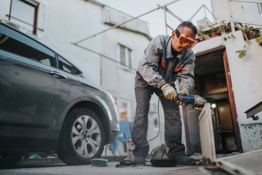 A focused construction worker wearing safety goggles and gloves operates an angle grinder to cut a wooden panel in a driveway area beside a parked car, sparks flying as power tools buzz.