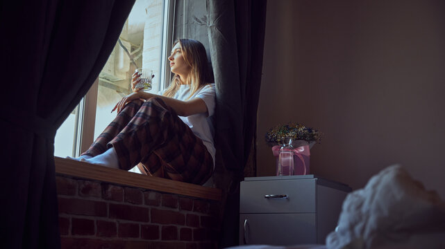 Young woman sitting on windowsill with drink in peaceful morning mood. Concept of mindfulness, lifestyle storytelling, calm routines, relaxation ads, and wellbeing promotional content.