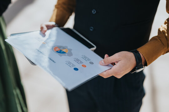 A business professional in smart casual attire presents a folder with charts in an urban environment, conveying professional collaboration, planning, and data presentation in a city context.