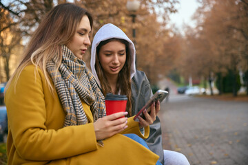 Two young women with coffee outdoors checking smartphone in autumn scenery. Concept of lifestyle friendship, social media visuals, youth communication, beverage branding, and outdoor storytelling.