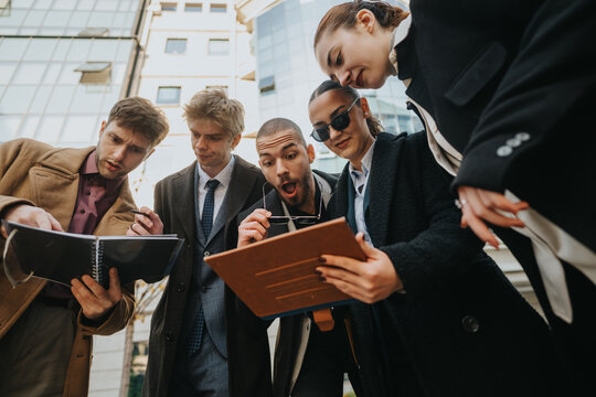 A group of young coworkers in smart casuals stands outside a glass office building, examining folders and a tablet, sharing ideas and collaborating in a dynamic urban environment. - Powered by Adobe