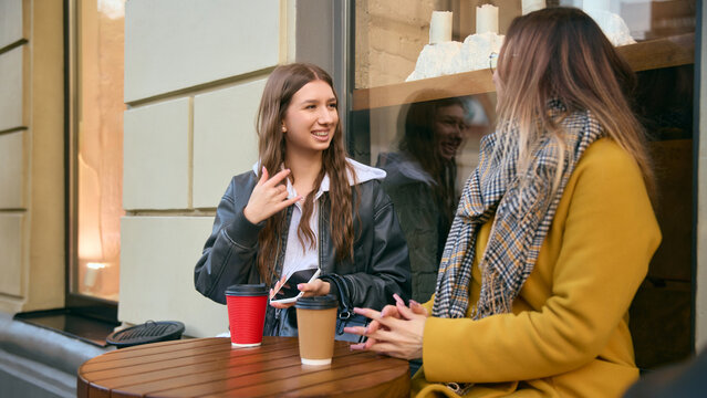 Young women talking over coffee at outdoor cafe table in cozy autumn street. Concept of lifestyle communication, beverage culture, friendship, socializing, and relaxed youth interaction.