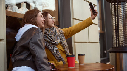 Two young women taking selfie at outdoor cafe table with takeaway coffee cups. Concept of lifestyle social media culture, beverage enjoyment, friendship, youth communication, and urban leisure moments
