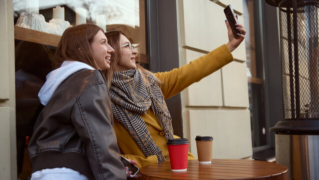 Two young women taking selfie at outdoor cafe table with takeaway coffee cups. Concept of lifestyle social media culture, beverage enjoyment, friendship, youth communication, and urban leisure moments