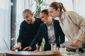 Three young coworkers in smart and formal attire gather around a laptop, sharing ideas and analyzing data in a bright open-plan office. They show teamwork, focus, and professional collaboration.