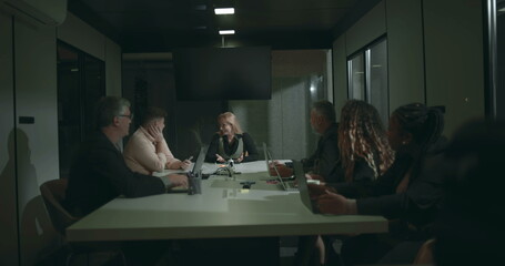 Diverse business team in serious discussion around boardroom table during meeting, using laptops and tablets, seated across modern office conference room