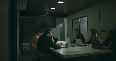Diverse business team in serious discussion around boardroom table during meeting, using laptops...