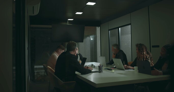 Diverse business team in serious discussion around boardroom table during meeting, using laptops and tablets, seated across modern office conference room - Powered by Adobe