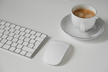 A workspace with a minimalist white keyboard and mouse, a white cup of coffee on a saucer. Ideal for topics: morning, freelancing, technology, productivity.