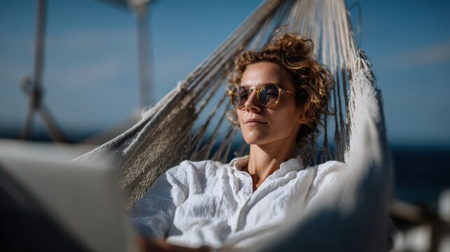 A relaxed woman reclines in a hammock by the sea, enjoying a tranquil moment while working on her laptop, epitomizing leisure, serenity, and the joy of remote work.