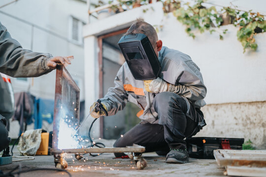 A skilled welder crouches at an outdoor construction area, welding a metal frame while sparks fly. Protective gear and gloves emphasize safety in a busy, hands-on metalwork setting.
