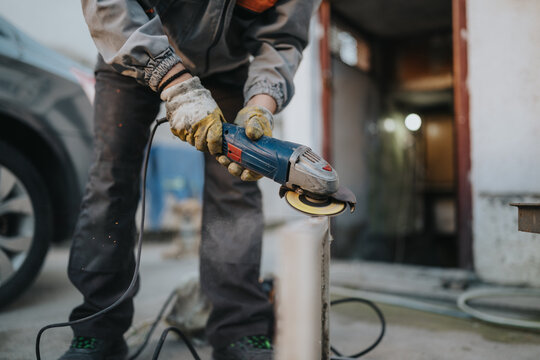 Construction worker wearing gloves operates a grinder to smooth metal, producing bright sparks in a busy outdoor workshop. The power tool and cord emphasize manual labor and focused craftsmanship.