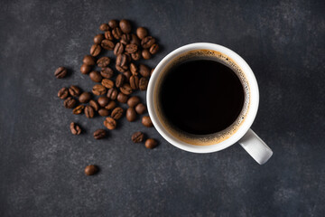 Black coffee in white cup on dark stone background, roasted coffee beans shattered around the cup. Top view