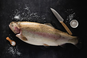 Fresh trout fish, sea salt and kitchen knife on black slate background. Cooking fish, recipe menu background