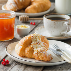 Tasty almond croissant, cup of black coffee, jam and butter on wooden table background