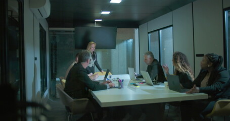 Female executive walking into boardroom to address coworkers during business meeting with diverse team in modern office at night