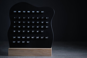 Modern black perpetual calendar on wooden base stands against dark background, featuring round buttons for days and months.
