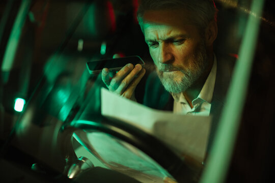Mature man looking concerned using phone and paperwork in car at night