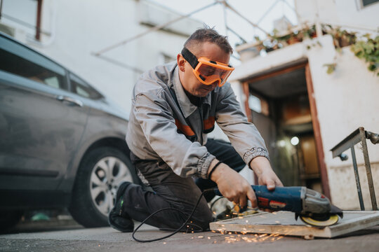 A construction worker crouches on a driveway, cutting a wooden board with an angle grinder. Sparks spray as he concentrates, wearing protective gear in an outdoor work scene with a car nearby.