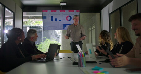 Diverse team of professionals attending meeting as manager points to presentation screen showing data charts in bright office conference room
